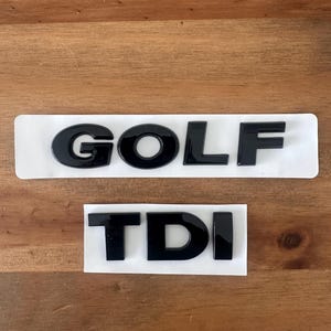 May include: Black plastic "GOLF" and "TDI" car badges on white backing. The letters are glossy. The badges are displayed on a wooden surface.