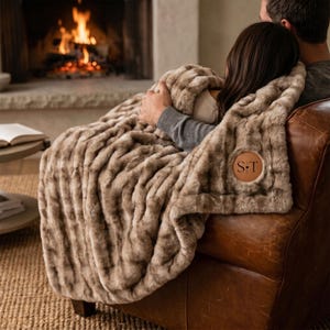 May include: A plush, textured faux fur throw blanket in a warm tan color, draped over a brown leather armchair. The blanket features a circular tan leather patch on the corner with the embossed letters S and T separated by a small heart symbol.