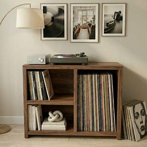 Puede incluir: Un mueble de discos de madera con tocadiscos, amplificador y arte enmarcado. El mueble está lleno de discos de vinilo. A la izquierda hay una lámpara de pie dorada con una pantalla beige.