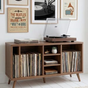May include: A wooden record player console in a mid-century modern style, with a warm brown finish. The unit has multiple compartments filled with vinyl records. A turntable with a clear cover is on top, with framed vintage music posters on the wall.