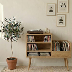 Puede incluir: Consola de tocadiscos de madera clara con almacenamiento de discos integrado. La unidad cuenta con tres estantes y un cajón, llenos de discos de vinilo. A la izquierda hay un pequeño árbol en una maceta de terracota y arte enmarcado en la pared.
