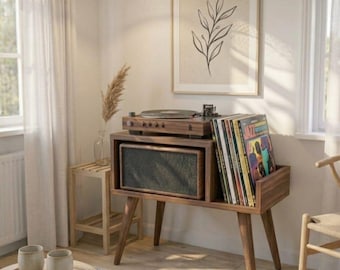 May include: A mid-century modern record player console in a living room setting. The wooden console features a turntable, speaker, and record storage. A framed botanical print hangs above. The room is decorated in neutral tones with natural light.