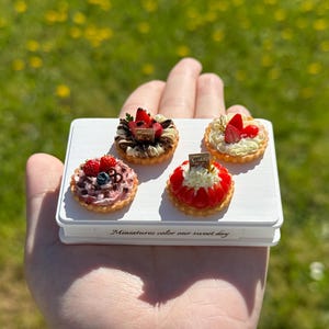 Puede incluir: Miniaturas de tartaletas de frutas, con fresas y arándanos, presentadas en una bandeja rectangular blanca. La bandeja tiene el texto "Miniatures color our sweet day". El fondo es hierba verde borrosa.