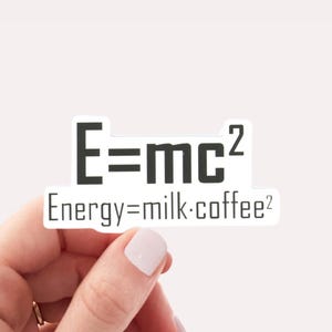 May include: A white sticker with black text that reads "E=mc² Energy=milk.coffee²". A hand is holding the sticker.