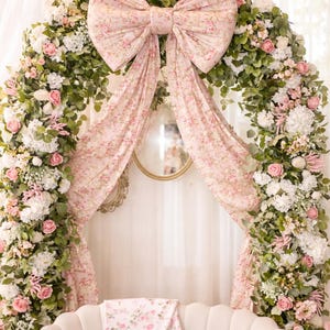 May include: A floral archway with a large pink bow and draping fabric. The arch is decorated with white and pink flowers and greenery. A white sofa with a floral blanket is in front of the arch.