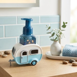 May include: A novelty soap dispenser shaped like a vintage camper trailer. The dispenser is white and light blue with a dark blue pump. The dispenser sits on a wooden surface, with a small vase and towel in the background.