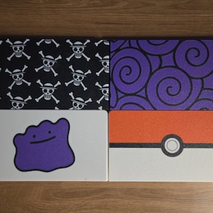 May include: Four rectangular mouse pads with different designs. One features a black background with white skull and crossbones, another has purple swirls, a third has a purple Ditto character, and the last is a red and white Pokeball.