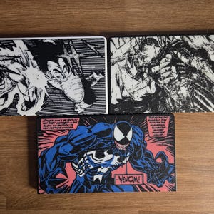 Puede incluir: Tres posavasos rectangulares con arte de cómic. Uno presenta una ilustración en blanco y negro de una escena de batalla. Otro representa a un personaje en posición de combate. El tercer posavasos muestra a Venom con la palabra "Venom!"