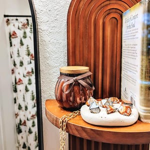 May include: A brown glass jar with a wooden lid and a brown ribbon sits on a wooden shelf. A small white dish holds several dice. A gold chain hangs down. A book titled "Words of Comfort for the Soul" is also on the shelf.