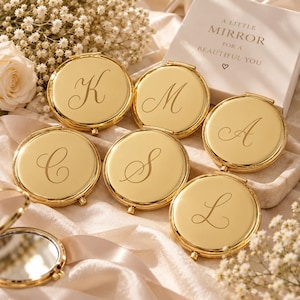May include: A collection of round, gold-toned metal compact mirrors, each engraved with a single cursive letter. A white box is present with the text A LITTLE MIRROR FOR A BEAUTIFUL YOU and a small heart symbol printed on the front surface.
