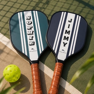 Peut inclure: Deux raquettes de pickleball avec des poign&eacute;es aspect cuir marron et des bords noirs. Une raquette est vert fonc&eacute; avec des rayures blanches et le nom SOPHIE, l'autre est bleu marine avec des rayures blanches et le nom JIMMY. Logo Tommy Bahama et balle jaune.
