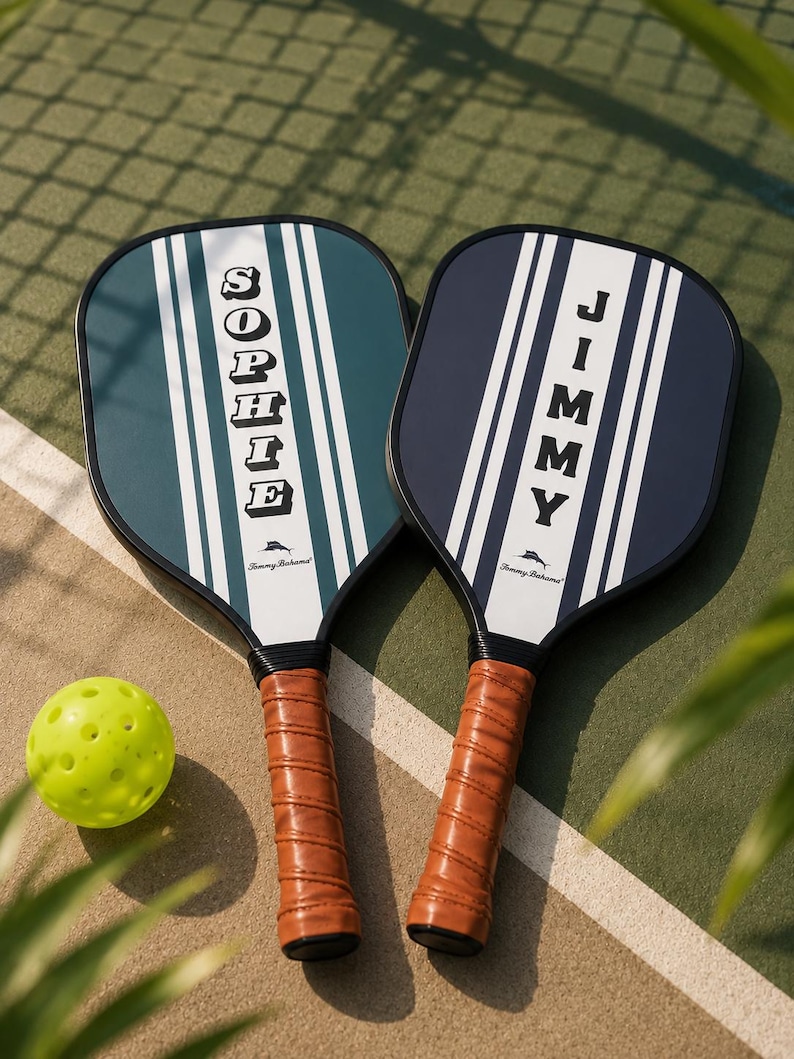 Peut inclure: Deux raquettes de pickleball avec des poign&eacute;es aspect cuir marron et des bords noirs. Une raquette est vert fonc&eacute; avec des rayures blanches et le nom SOPHIE, l'autre est bleu marine avec des rayures blanches et le nom JIMMY. Logo Tommy Bahama et balle jaune.