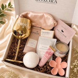 May include: A gift box labeled BOXORA containing a gold wine glass with the name Angel, a pink silk sleep mask, a Moonstruck milk chocolate bar, a lavender soap bar, a Peauamie rose eye mask, a bath bomb, a tube of hand cream, a candle, and a flower-shaped item.