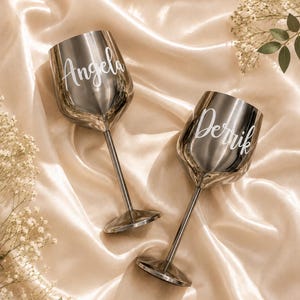 May include: Two metallic silver wine glasses with long stems and round bases. Each glass features white cursive lettering, with one glass displaying the name Angela and the other glass displaying the name Derrik, set against a soft, draped fabric.