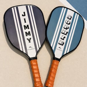 Peut inclure: Deux raquettes de pickleball avec des bords noirs et des poign&eacute;es envelopp&eacute;es de cuir orange. La raquette de gauche est bleu marine fonc&eacute; avec des rayures blanches et le nom JIMMY. Celle de droite est bleu clair avec le nom SOPHIE. Logo Tommy Bahama.