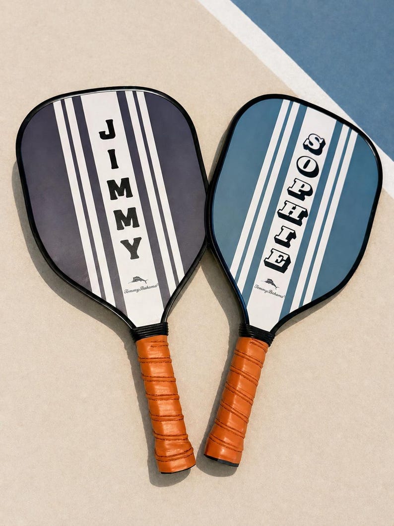 Peut inclure: Deux raquettes de pickleball avec des bords noirs et des poign&eacute;es envelopp&eacute;es de cuir orange. La raquette de gauche est bleu marine fonc&eacute; avec des rayures blanches et le nom JIMMY. Celle de droite est bleu clair avec le nom SOPHIE. Logo Tommy Bahama.