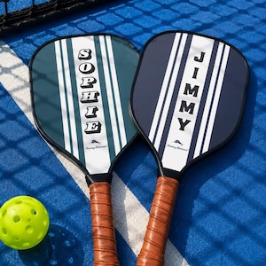 Peut inclure: Deux raquettes de pickleball avec des poign&eacute;es envelopp&eacute;es de cuir marron. La raquette de gauche est bleu sarcelle avec des rayures blanches et le nom SOPHIE. Celle de droite est bleu marine avec des rayures blanches et le nom JIMMY. Une balle jaune.