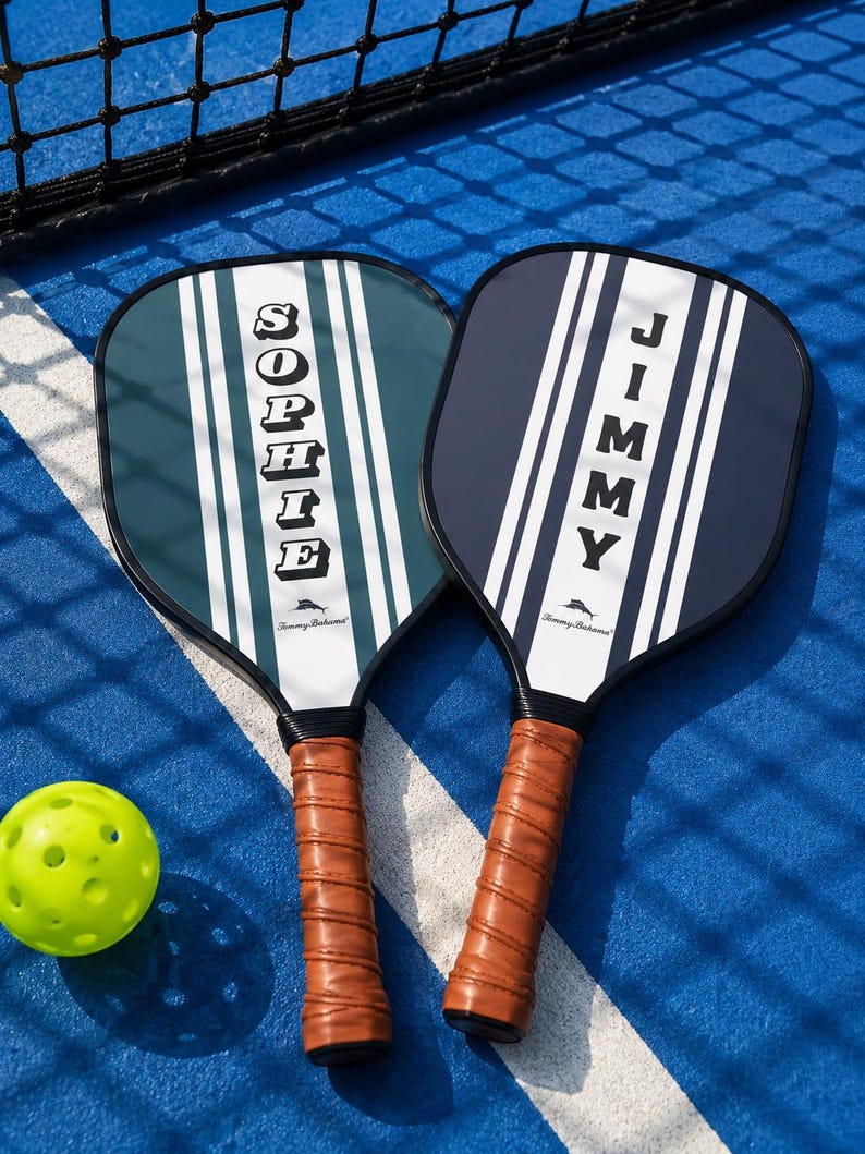 Peut inclure: Deux raquettes de pickleball avec des poign&eacute;es envelopp&eacute;es de cuir marron. La raquette de gauche est bleu sarcelle avec des rayures blanches et le nom SOPHIE. Celle de droite est bleu marine avec des rayures blanches et le nom JIMMY. Une balle jaune.