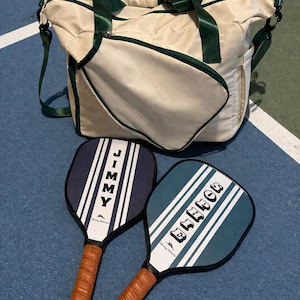 Peut inclure: Un sac de sport cr&egrave;me avec des sangles et des bordures vert fonc&eacute;, accompagn&eacute; de deux raquettes de pickleball. Une raquette est bleu marine avec le nom JIMMY, et l'autre est bleu sarcelle avec le nom DONNA. Les deux ont des rayures blanches et des poign&eacute;es en cuir marron.