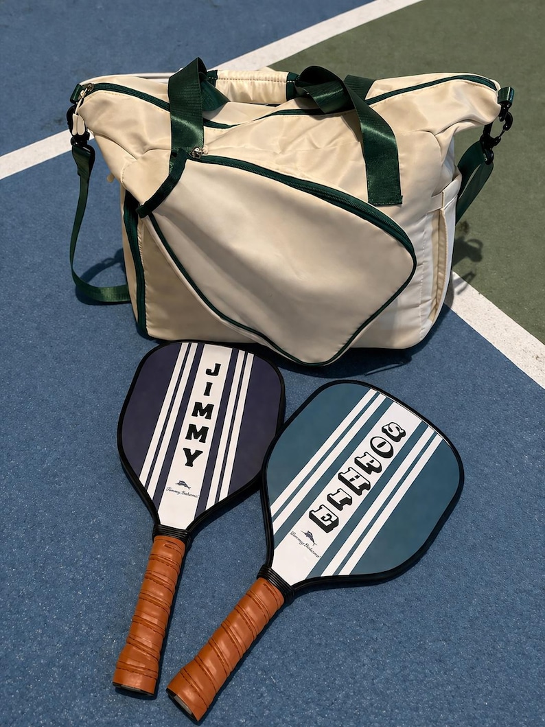 Peut inclure: Un sac de sport cr&egrave;me avec des sangles et des bordures vert fonc&eacute;, accompagn&eacute; de deux raquettes de pickleball. Une raquette est bleu marine avec le nom JIMMY, et l'autre est bleu sarcelle avec le nom DONNA. Les deux ont des rayures blanches et des poign&eacute;es en cuir marron.