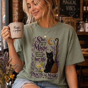 May include: A sage green t-shirt featuring a graphic print of a black cat, a broomstick, a cocktail, and lavender sprigs with the text: She had the gift of Magic, and it was this very gift that saved her life, PRACTICAL MAGIC. A beige mug reads Magic in progress.