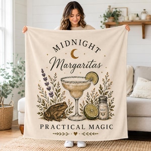 May include: A cream-colored throw blanket featuring a central illustration of a margarita in a glass, a toad, a salt shaker, and sprigs of lavender. The text reads Midnight Margaritas and Practical Magic, accented by a crescent moon and stars.