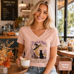 Peut inclure: Un t-shirt col en V couleur pêche chiné avec une impression graphique représentant un croissant de lune, un chapeau de sorcière, un balai et un chat noir. Le texte indique Practical Magic et Fall in love whenever you can, entouré de fleurs.