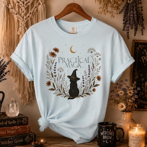 May include: A light blue t-shirt featuring a graphic of a black cat wearing a witch hat, surrounded by a floral wreath and a crescent moon. The text "PRACTICAL MAGIC" is printed in a serif font above the cat. The shirt has short sleeves.