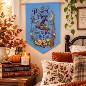 Peut inclure: Une bannière murale en tissu bleu clair ornée d'un chapeau de sorcière violet foncé, d'un balai, d'un livre ouvert et d'illustrations florales. Le texte indique "Practical Magic" et "There's No Devil In The Craft" en écriture décorative.