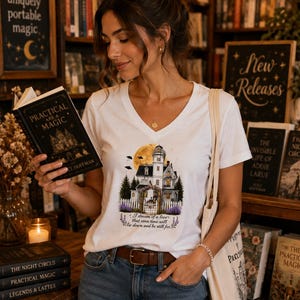 Peut inclure: Un t-shirt blanc à col en V avec une impression graphique représentant une maison victorienne, une grande lune jaune, des pins et une clôture blanche. Le texte sous la maison indique, I dream of a love that even time will lie down and be still for.