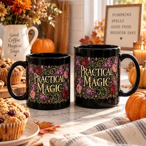 May include: Two black ceramic mugs featuring the text "PRACTICAL MAGIC" in gold lettering, surrounded by a detailed floral design of red roses, purple flowers, and green vines. The mugs have a glossy finish and a standard handle on the side.