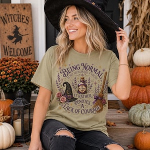 May include: A khaki t-shirt featuring a graphic print with the text "Being normal is not necessarily a virtue, it rather denotes a lack of courage." The design includes a witch hat, a potion bottle, a broom, and floral accents in purple and pink.