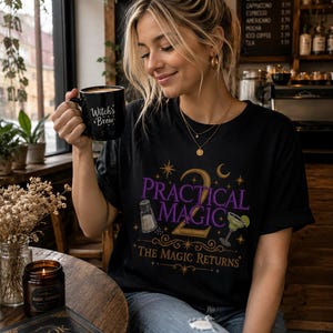 May include: A black t-shirt featuring a graphic print with the text Practical Magic 2 and The Magic Returns in purple and gold. The design includes illustrations of a salt shaker, a margarita glass, stars, and a crescent moon. The shirt is worn with blue jeans.
