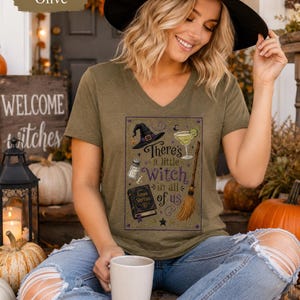 May include: A heather olive V-neck t-shirt featuring a graphic print of a witch hat, a cocktail, a salt shaker, a broom, and a book titled Amas Veritas, with the text There's a little witch in all of us, surrounded by small stars and moons.