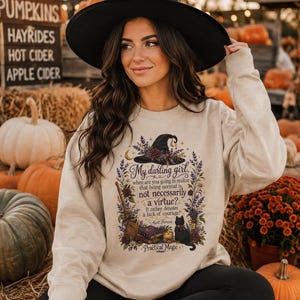 May include: A sand-colored crewneck sweatshirt featuring a graphic print of a witch hat, a broom, a black cat, and floral elements. The text reads: My darling girl, when are you going to realize that being normal is not necessarily a virtue? It rather denotes a lack of courage! - Aunt Frances. Practical Magic.