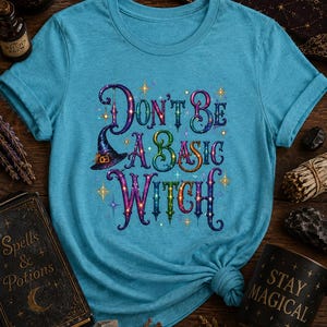 May include: A teal short-sleeved t-shirt featuring a colorful graphic print with the text "Don't Be A Basic Witch" in a stylized font. The design includes a purple witch hat and small star accents in various colors, centered on the chest.