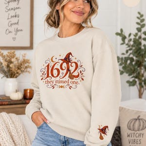 May include: A sand-colored crewneck sweatshirt featuring a graphic print with the text 1692 they missed one, a witch hat, and a broomstick. The design includes autumn leaves and stars. A smaller matching witch hat and broomstick graphic is on the left sleeve.