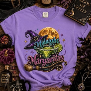 May include: A light purple t-shirt featuring a graphic print of a witch hat, a full moon, and a margarita glass with a lime slice. The text reads Midnight Margaritas. The design includes stars, a broomstick, and glittery accents in shades of green, purple, and gold.
