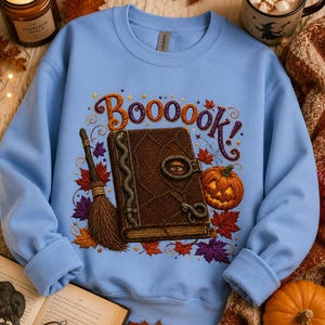May include: A light blue crewneck sweatshirt featuring a graphic print of a brown spellbook with a snake and an eye on the cover, a broomstick, a jack-o'-lantern, and autumn leaves. The text "Boooook!" is printed in purple above the book.