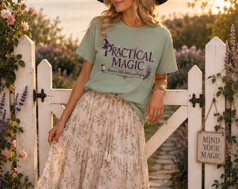 May include: A sage green t-shirt featuring the text "PRACTICAL MAGIC" and "there's a little witch in all of us" with a witch hat and lavender graphic, paired with a long, tiered, cream-colored skirt with a delicate pink and green floral print.