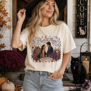 May include: A cream-colored short-sleeve t-shirt featuring a graphic print of a couple, a house, a black cat, a candle, and a book. The text reads "Practical Magic" and "I wished for you too" surrounded by a colorful floral and leaf border.