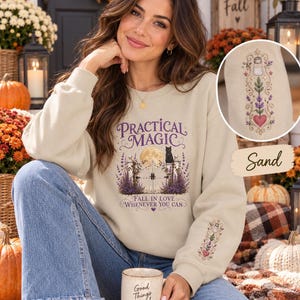 May include: A sand-colored crewneck sweatshirt featuring a graphic print with the text Practical Magic, Fall in Love Whenever You Can, a full moon, a black cat, and iron gates. The sleeve includes an embroidered design of a salt shaker and heart.