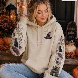 May include: A sand-colored hooded sweatshirt featuring a graphic of a purple witch hat and the text Practical Magic on the chest. The sleeves are decorated with illustrations of black cats, cocktails, umbrellas, flowers, and a large house.
