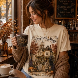 May include: A cream-colored t-shirt featuring a graphic of a large house with a white picket fence, pumpkins, and a black cat. The text reads Practical Magic and There's a Little Witch in All of Us. The shirt is worn with a brown knit cardigan.