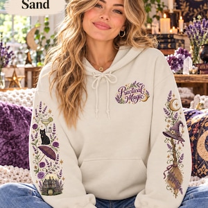 May include: A sand-colored hooded sweatshirt featuring the text Practical Magic on the chest. The sleeves are decorated with detailed illustrations of lavender, a black cat, a cocktail, a parasol, a house, a witch hat, and a broomstick.