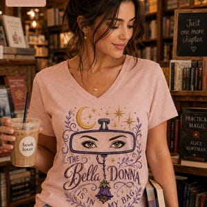 Peut inclure: Un t-shirt à col en V couleur pêche chiné avec un motif de rétroviseur aux yeux, un croissant de lune, des étoiles et des vignes florales. Le texte indique "THE BELLA DONNA IS IN MY BAG". Un petit encadré affiche "Heather Peach".