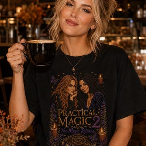 May include: A black t-shirt featuring a graphic print of two women with long hair, surrounded by candles, purple butterflies, and floral accents. Below them, the text reads Practical Magic 2 The Magic Returns above an illustration of a spellbook.