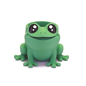 Könnte beinhalten: Ein grüner, froschförmiger Pflanztopf mit großen, ausdrucksstarken Augen und einem lächelnden Mund. Der Pflanztopf hat eine ausgehöhlte Oberseite, um kleine Pflanzen oder dekorative Gegenstände aufzunehmen. Die Füße des Frosches sind gespreizt, was ihm ein verspieltes Aussehen verleiht.