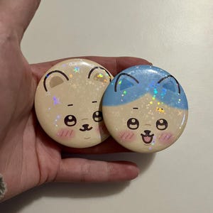 May include: Two round, cartoon-style pinback buttons. One is tan with a light blue top, the other is tan. Both have a cute bear face design with black eyes, a nose, and a smiling mouth. The buttons have a glittery, holographic effect.