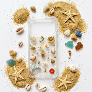Puede incluir: Funda transparente para teléfono decorada con varios abalorios, incluyendo un caballito de mar, una estrella de mar y una flor roja. La funda está rodeada de arena, conchas marinas y pequeñas piedras, creando una estética de playa.